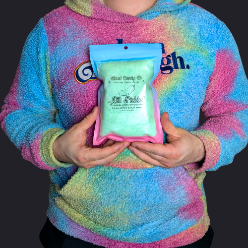 A person wearing a pastel tie-dye fleece hoodie holds a clear stand-up pouch filled with dill pickle cotton candy. The pouch has a blue top and pink bottom trim, with visible branding for Cloud Candy Co.