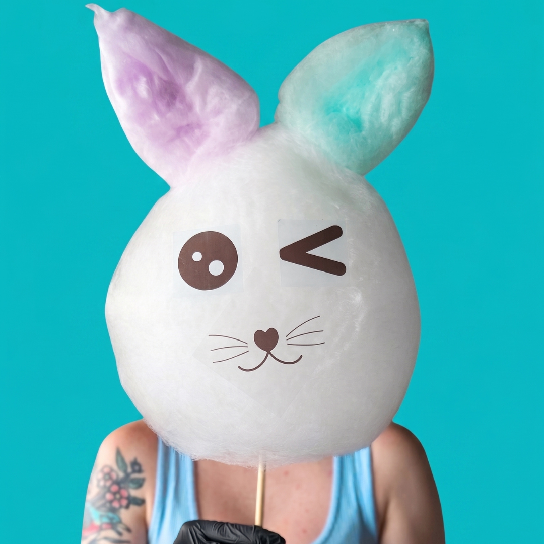 A person wearing black gloves holds a large white bunny-shaped cotton candy on a stick against a bright turquoise background. The bunny has pastel purple and mint blue ears, a winking face, and a small heart nose.