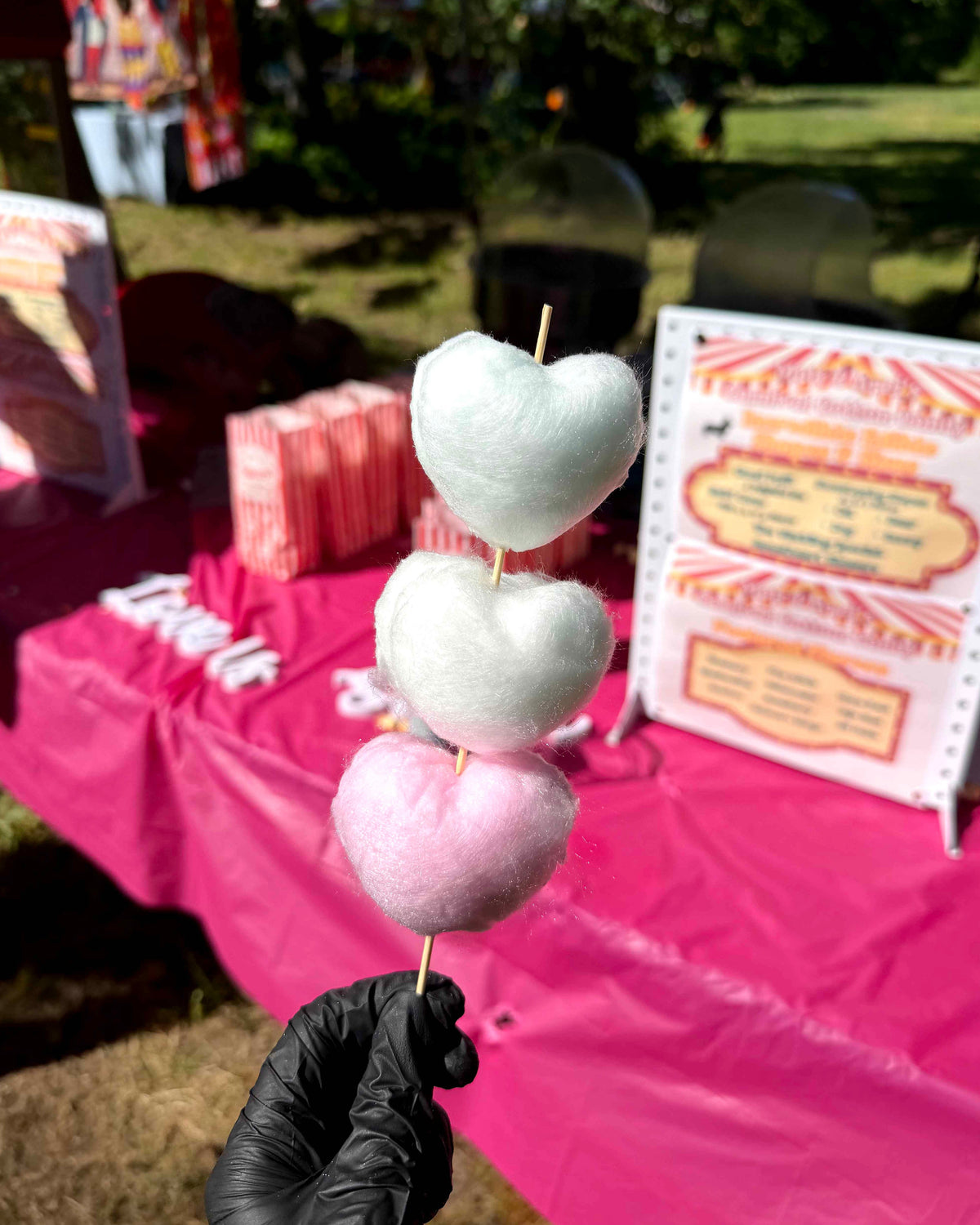 A hand wearing a black glove holds a skewer of three heart-shaped cotton candy pieces in pink and white, with a cotton candy booth in an outdoor park setting in the background.