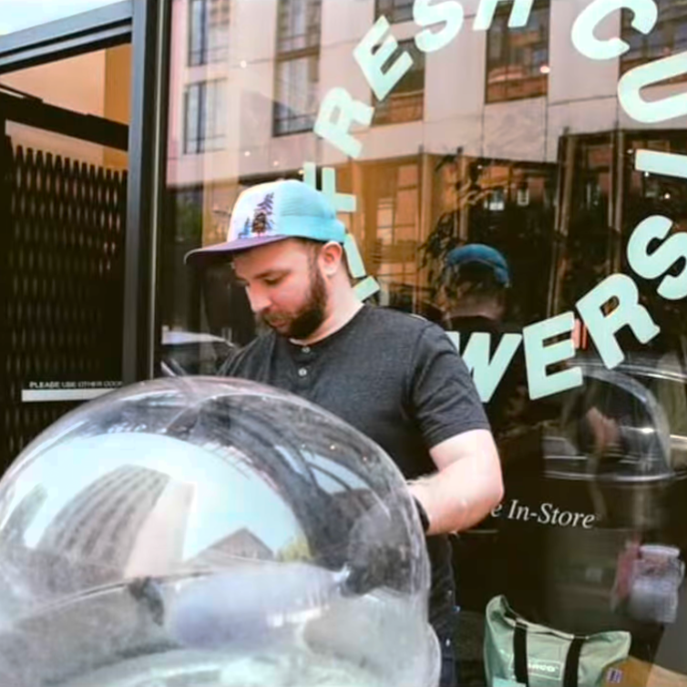 A man wearing a black t-shirt and a blue and white baseball cap stands in front of a glass storefront window with large green lettering that reads 'FRESH CUTS'. A clear plastic dome-like object is prominently visible in the foreground, reflecting the street scene.
