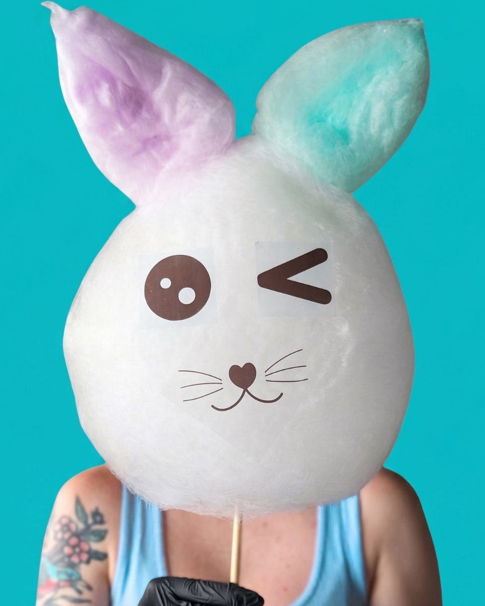 A person wearing black gloves holds a large white bunny-shaped cotton candy on a stick against a bright turquoise background. The bunny has pastel purple and mint blue ears, a winking face, and a small heart nose.