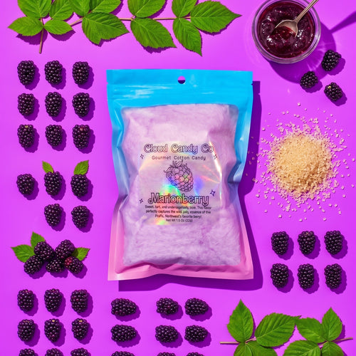 A bag of Cloud Candy Co. Marionberry cotton candy is displayed in a flat-lay composition on a bright magenta background, surrounded by blackberries, green leaves, a jar of marionberry jam, and sugar crystals.