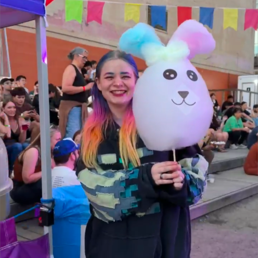 A smiling woman with rainbow-colored hair stands outdoors at what looks like a fair or event, holding a large white cotton candy bunny with pastel blue and pink ears. A vendor booth and seated crowd are visible in the background.