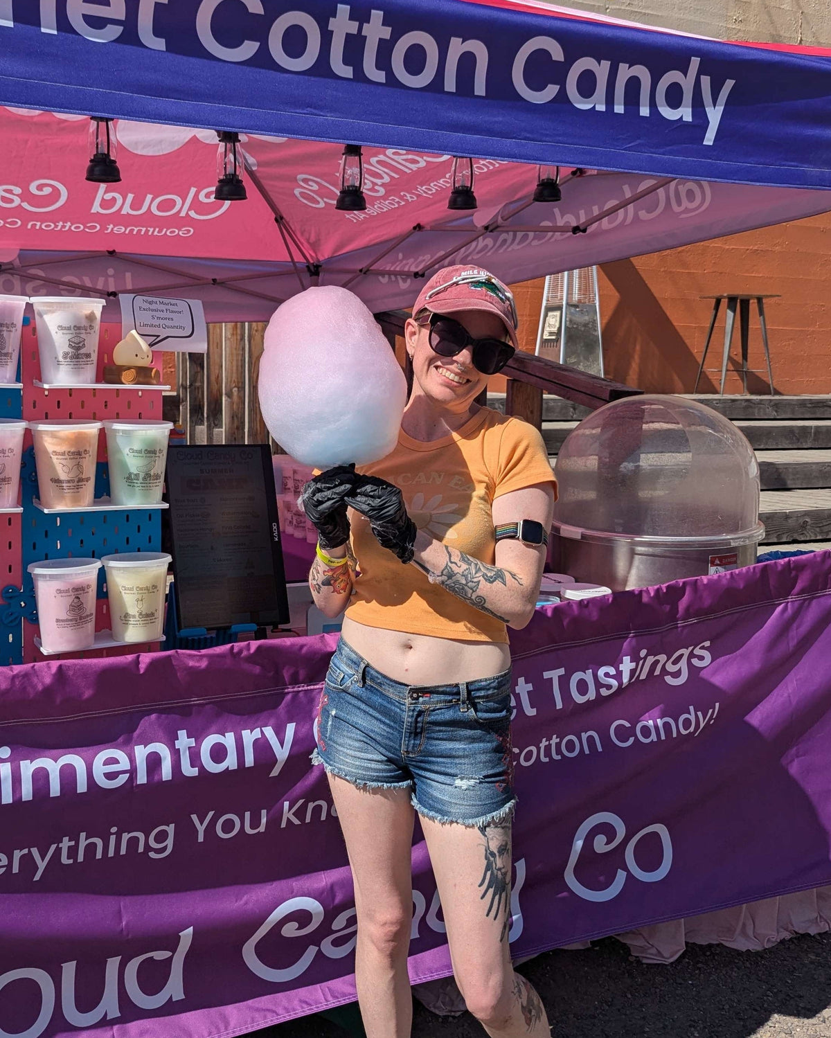 A woman stands outdoors in front of a cotton candy stand, holding a large pink and white cotton candy ball. The purple and pink canopy tent has visible branding and displays cotton candy cups and packaging.